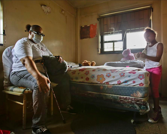 Wanda Coleman clutches her aching knee while watching television with her granddaughter, Jasmine Hodge, in her public housing apartment in New York. Coleman, who wears a mask for severe asthma, is about to be evicted, partly for her son's criminal activity, due to "One Strike and You're Out" policies and will have to go to a homeless shelter.