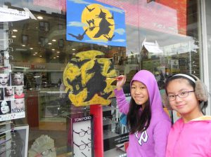 Sara Dang (left) and Jessica Chin paint a witch on the window of Lens Lab Express. Eagle photos by Paula Katinas