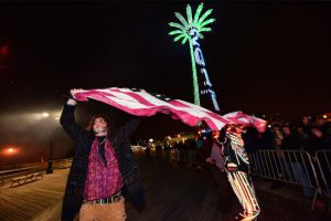 A visitor waves a U.S. flag after midnight on the Coney Island Boardwalk with the illuminated Parachute Jump in the background. Eagle photos by Andy Katz