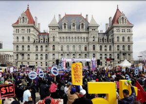 In this 2016 file photo, the state Capitol provides a backdrop as supporters of a $15 minimum wage rally at the Empire State Plaza in Albany. Millions of workers across the U.S. saw their pay increase as 19 states bump up their minimum wages as the new year begins. New York state is taking a regional approach, with the wage rising to $11 in New York City. AP Photo/Mike Groll, File