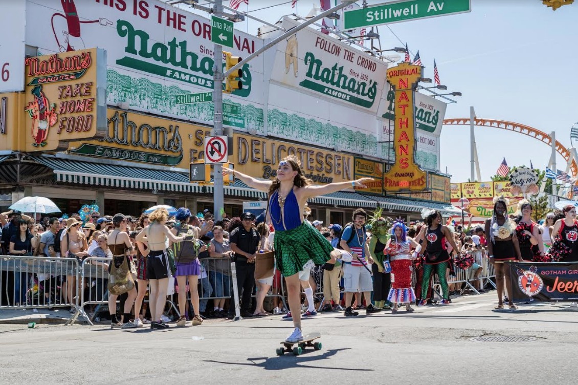 A woman skateboards past a crowd in front of the iconic Nathan’s Famous hot dog restaurant.