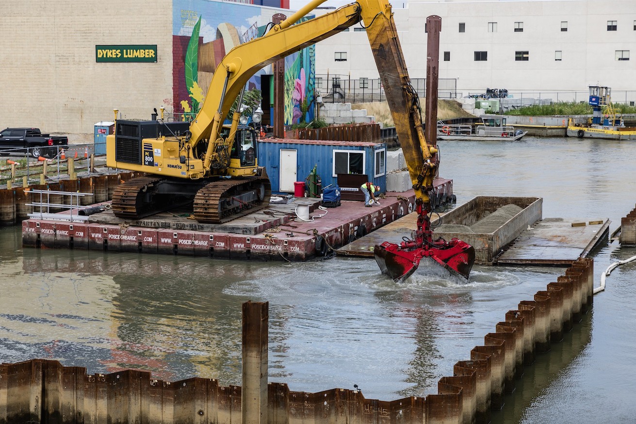 A crane operator floating on a barge on the Gowanus Canal lifts the machine’s arm out of the water after dropping sand to the bottom of the canal.
