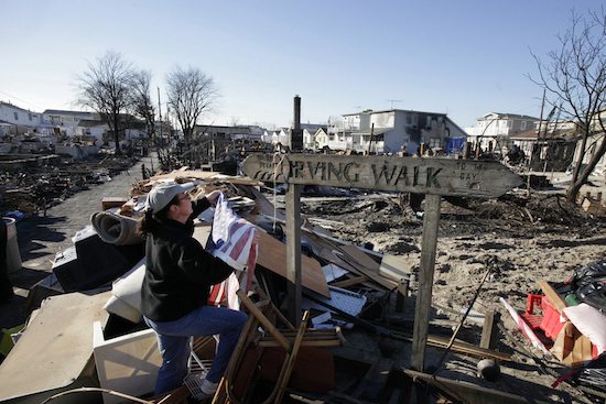 In this Nov. 14, 2012 photo, Louise McCarthy places an American flag on a street sign for Irving Walk in the Breezy Point neighborhood of Queens, N.Y. The sign survived a fire that swept through the seaside community during Superstorm Sandy two weeks earlier. Six years later, most residents have stayed. Homes have been restored, and Sandy led to a new ferry service and a rebuilt boardwalk. AP Photo/Mark Lennihan