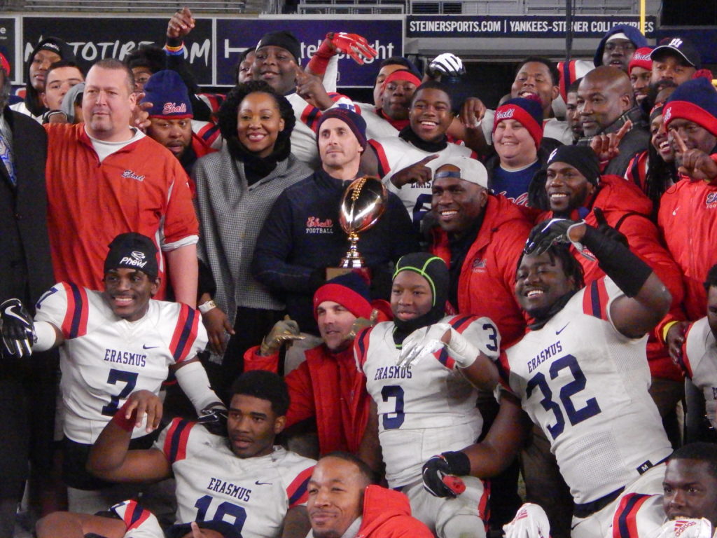 Holding the PSAL 2018 Championship Trophy in the center of the center of the pack, Erasmus Coach Danny Landberg along with his team celebrates their 34-7 win over South Shore at Yankee Stadium. Photo by Jim Dolan