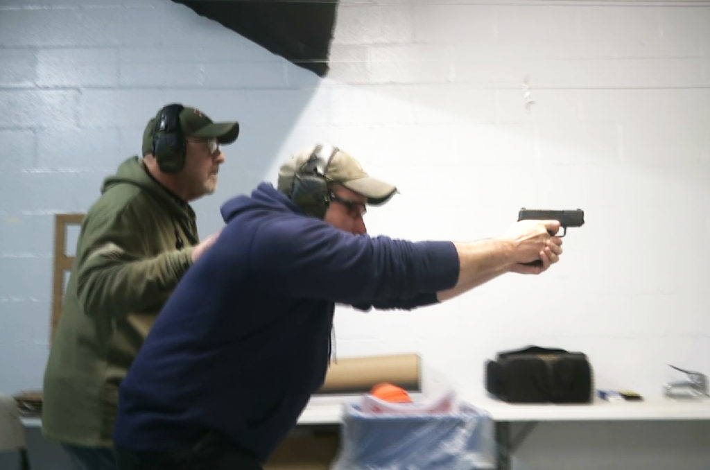 In this photo taken from a video shot on Nov. 28, 2018, Mike Carnevale places his hand on the back of Mark Hennesey while instructing him at the American Tactical Systems’ indoor range in Green Island, New York. The application process for handgun licenses would be expanded under a bill before the New York state Legislature. The bill would require handgun applicants to turn over log-in information so investigators could look at three years’ worth of Facebook, Snapchat, Twitter and Instagram postings. Google, Yahoo and Bing searches over the previous year also would be checked. (AP Photo/Michael Hill)