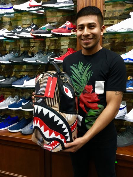 Gary Vergara, a manager at Es La Vida shoe store, poses with a Sprayground brand backpack that looks like a shark. Eagle photo by Lore Croghan