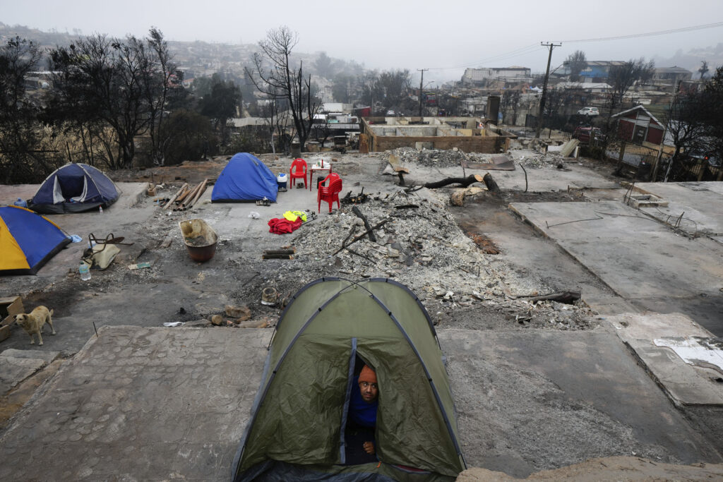 Chile — Nature’s revenge: More record-breaking wildfires: A man peers from the tent where he camped for the night at the spot where his home stood before it was destroyed by a forest fire in the Villa Independencia neighborhood of Vina del Mar, Chile, Tuesday, Feb. 6, 2024.Photo: Esteban Felix/AP