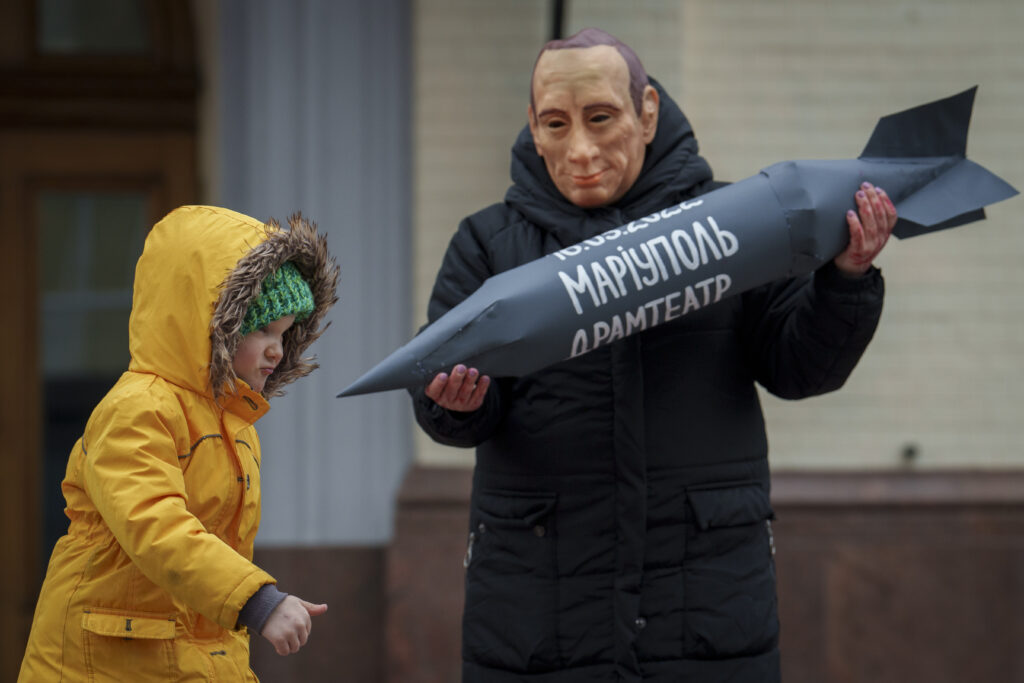 KYIV — Brightly-colored raincoat contrasts grimly costumed theater protest: A child runs past a person wearing a Vladimir Putin mask holding a cardboard missile with the words "Mariupol Drama Theatre" during a rally in Kyiv, Ukraine, Sunday, March 17, 2024. The rally, organized by the group "Azovstal Families" and attended by hundreds, called for renewed efforts for the release of Ukrainian servicemen from Mariupol, still in Russian captivity.Photo: Vadim Ghirda/AP