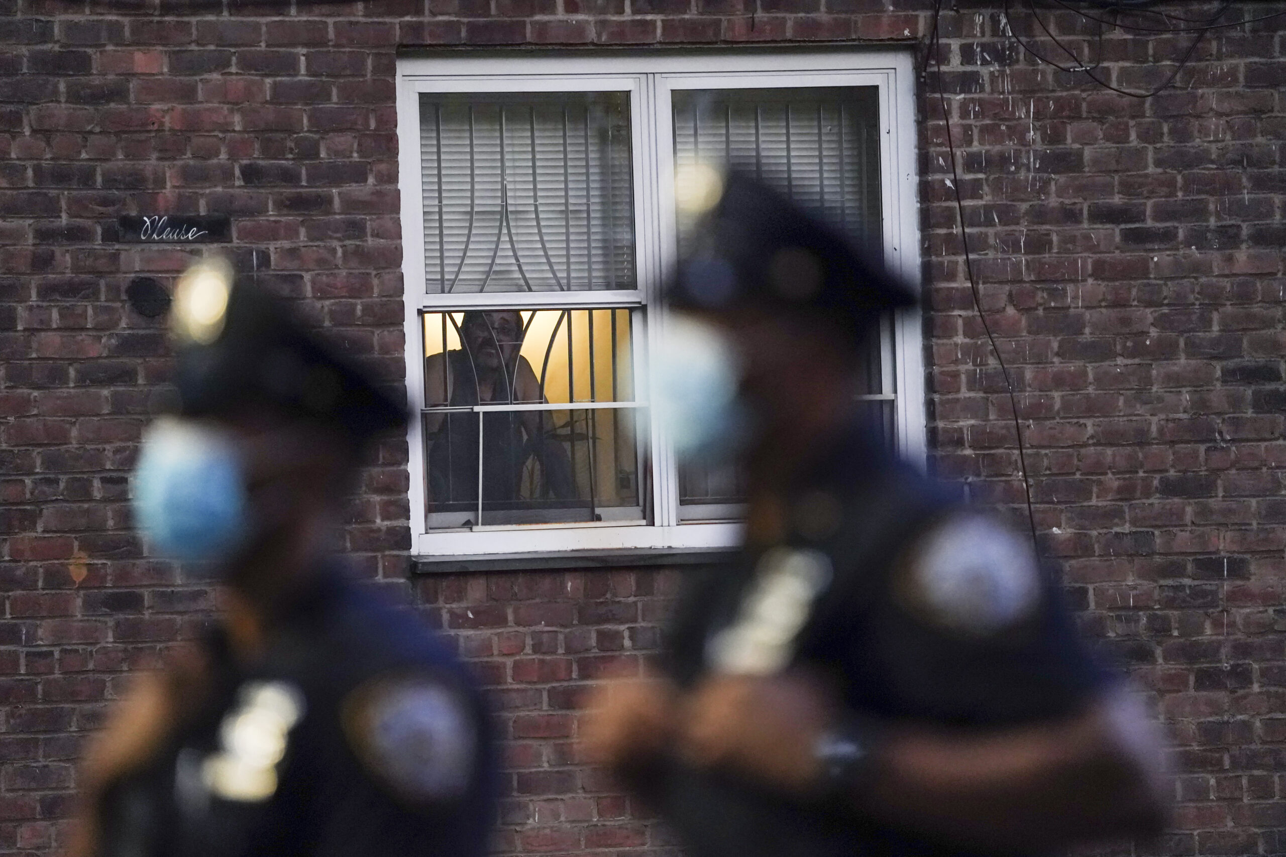 FILE - In this Aug. 18, 2020, file photo, a neighbor watches New York City Police Department officers work a crime scene where several young men were shot and wounded at the Ravenswood Houses in the Queens borough of New York. Heralded as the safest big city in America in recent years, New York City is closing out its bloodiest year in nearly a decade, grappling with a surge in homicides and a pandemic authorities say has helped fuel violence. (AP Photo/John Minchillo, File)