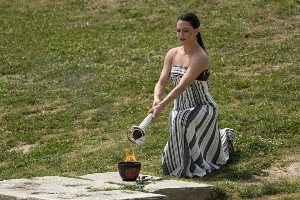 Actress Mary Mina, playing high priestess, lights a torch from a flame lit the day before during the official ceremony of the flame lighting for the Paris Olympics, at the Ancient Olympia site, Greece, Tuesday, April 16, 2024. Cloudy skies prevented the traditional lighting, when an actress dressed as an ancient Greek priestess uses the sun to ignite a silver torch. Instead, a backup flame was used that had been lit on the same spot Monday, during the final rehearsal. (AP Photo/Petros Giannakouris)