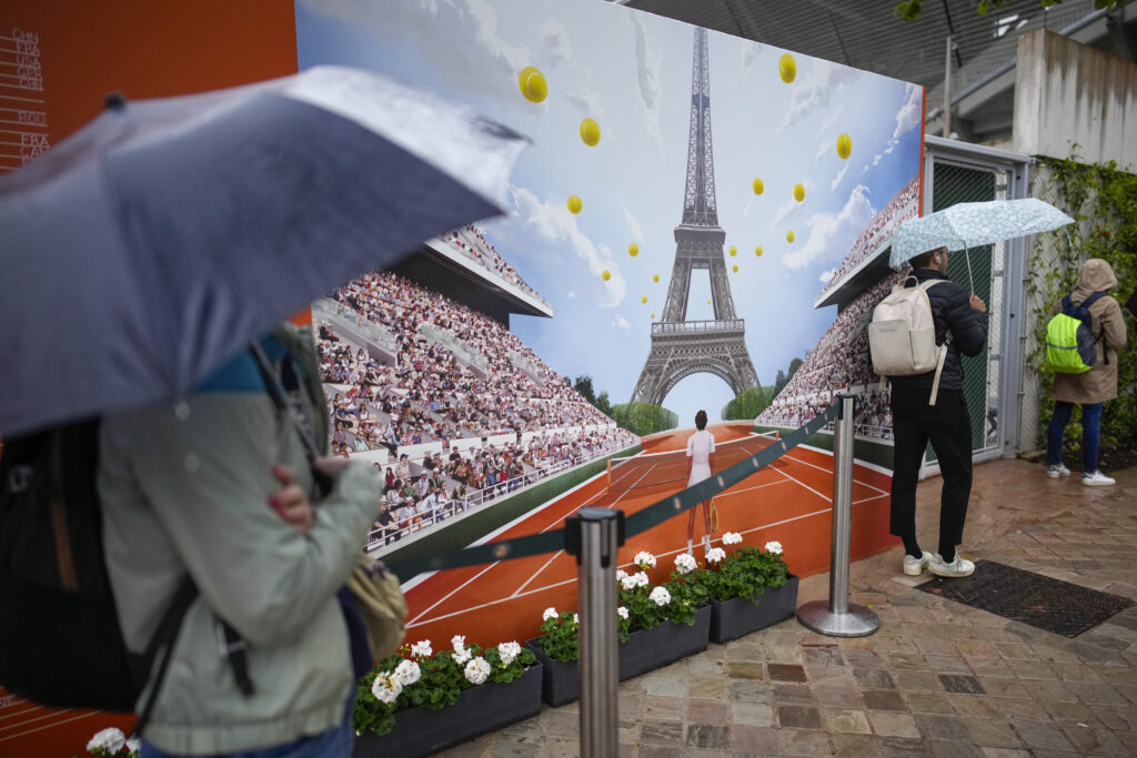 On a day where the weather was different from the illustration on the poster, tennis fans shield themselves from the rain which interrupted the majority of the second round matches of the French Open tennis tournament at the Roland Garros stadium in Paris, Wednesday, May 29, 2024. (AP Photo/Christophe Ena)