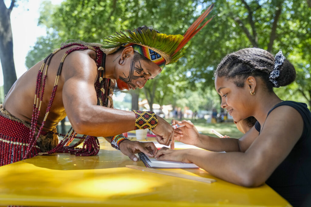 Txatxu Pataxo, left, of the Pataxo people of Bahia, Brazil, shows Eva Quiroz, 16, of Takoma Park, Md., how to draw a pattern traditional used in body painting, during the opening day of the Smithsonian Folklife Festival, Wednesday, June 26, 2024, on the National Mall in Washington. This year's festival theme is "Indigenous Voices of the Americas" and will run through July 1. "There was really no language barrier," says Quiroz, "because it's art and that's universal." (AP Photo/Jacquelyn Martin)