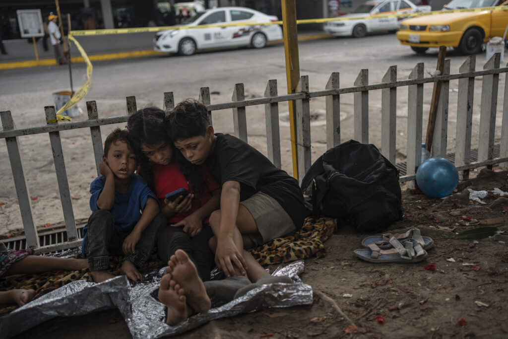 The Bolaños children, from left, Sebastian, Kamila and Miguel Angel, from Venezuela, watch videos on a mobile phone outside the bus terminal where they are living with their single mother Keilly and one other sibling, along with other migrants in Villahermosa, Mexico, Saturday, June 8, 2024. Their mother was captured in the northern state of Juarez, where she said she was beaten by the military in front of her children, loaded on a bus for two days, and left in Villahermosa. (AP Photo/Felix Marquez)