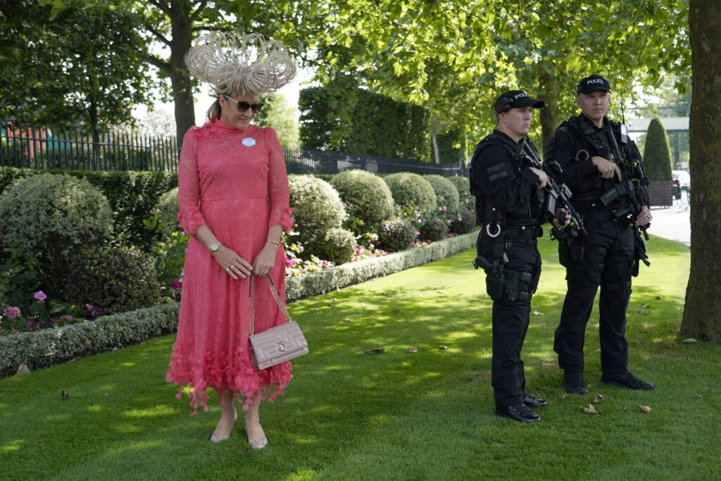 A racegoer pose for the media, as armed police stand guard near the main entrance on the first day of the Royal Ascot horse race meeting at Ascot, England, Tuesday, June 18, 2024. (AP Photo/Alberto Pezzali)