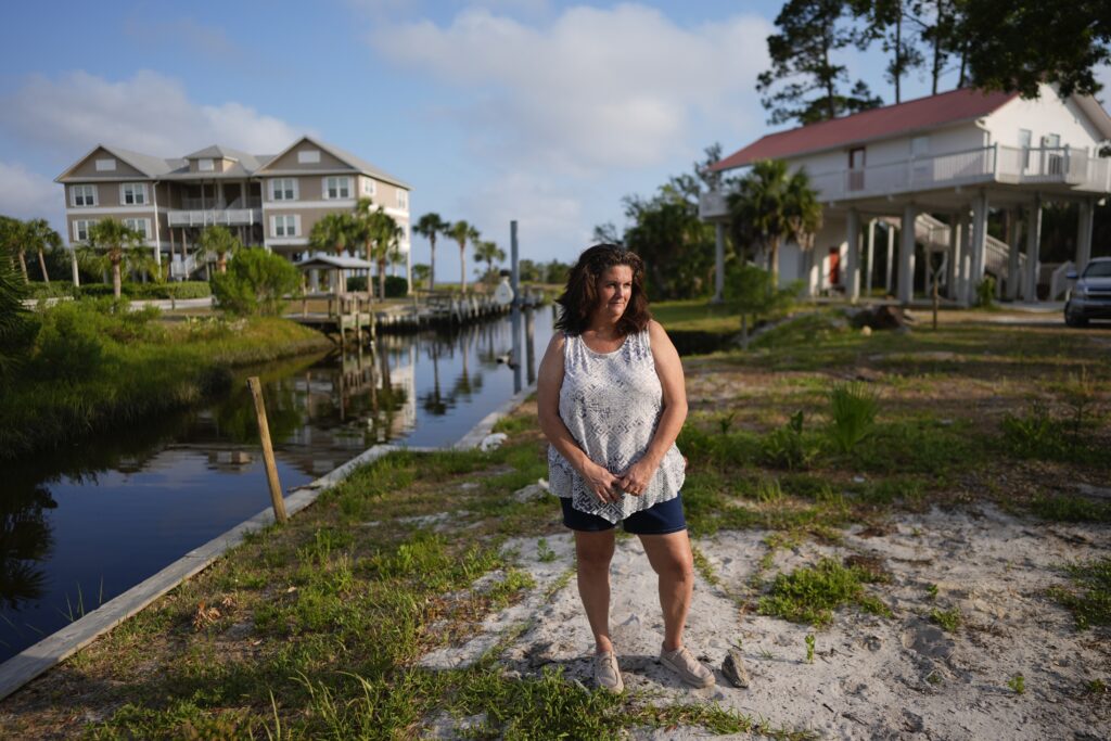 Lisa Bregenzer poses for a picture on the canal-side lot where her home stood until the passage of last year's Hurricane Idalia, Thursday, May 9, 2024, in Horseshoe Beach, Fla. In July, nearly a year after the storm that upended her and her husband's lives, Bregenzer has finally found hope. Her family was approved for a program that will help them build a new home, and if all goes as planned, they'll move into a stilted two-bedroom house on their lot by year's end. (AP Photo/Rebecca Blackwell)