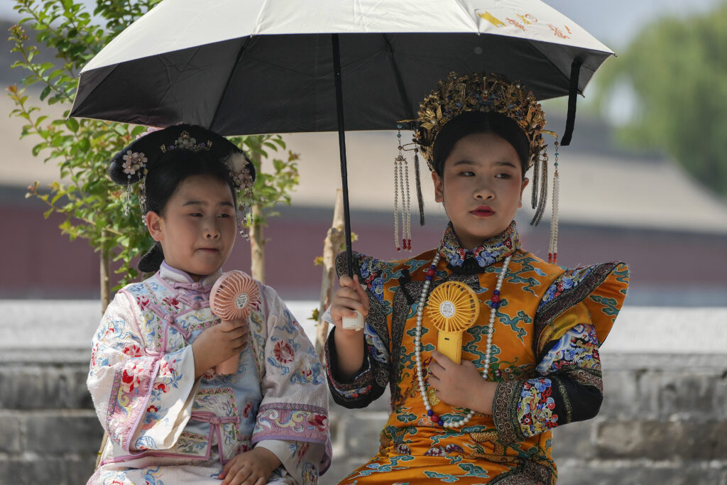 Children dressed in traditional costumes hold an umbrella and cool themselves with portable electric fan outside the Forbidden City on a sweltering day in Beijing, Monday, July 8, 2024. (AP Photo/Andy Wong)