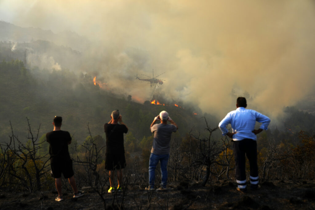 People watch and take photos of a helicopter dropping water on the flames during a third day of a wildfire, in Sofiana village, about 142 kilometers (88 miles) west of Athens, Greece, on Tuesday, Oct. 1, 2024. (AP Photo/Petros Giannakouris)