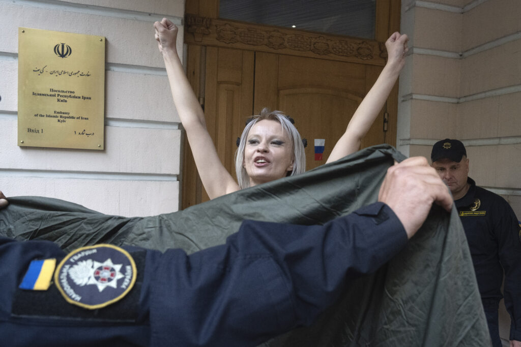 A police officer covers a topless activist of the Femen feminist group protesting against Russia-Iran military cooperation in front of the Iran Embassy in Kyiv, Ukraine, Friday, Oct. 4, 2024. (AP Photo/Efrem Lukatsky)