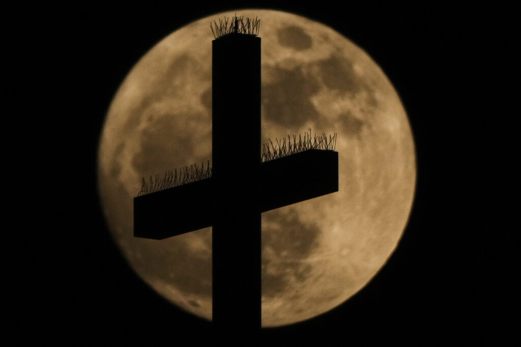 A cross atop the Cathedral of the Immaculate Conception Catholic Church is silhouetted against the rising full moon Sunday, May 11, 2025, in Kansas City, Mo.