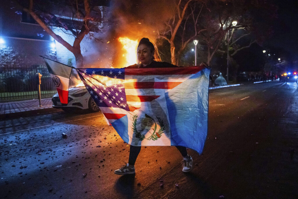A person displaying multiple flags walks past a burning car during protests over the Trump administration's immigration raids in Los Angeles, Monday, June 9, 2025.