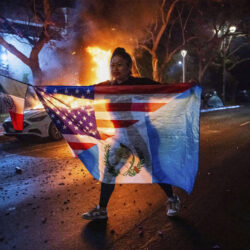 A person displaying multiple flags walks past a burning car during protests over the Trump administration's immigration raids in Los Angeles, Monday, June 9, 2025.
