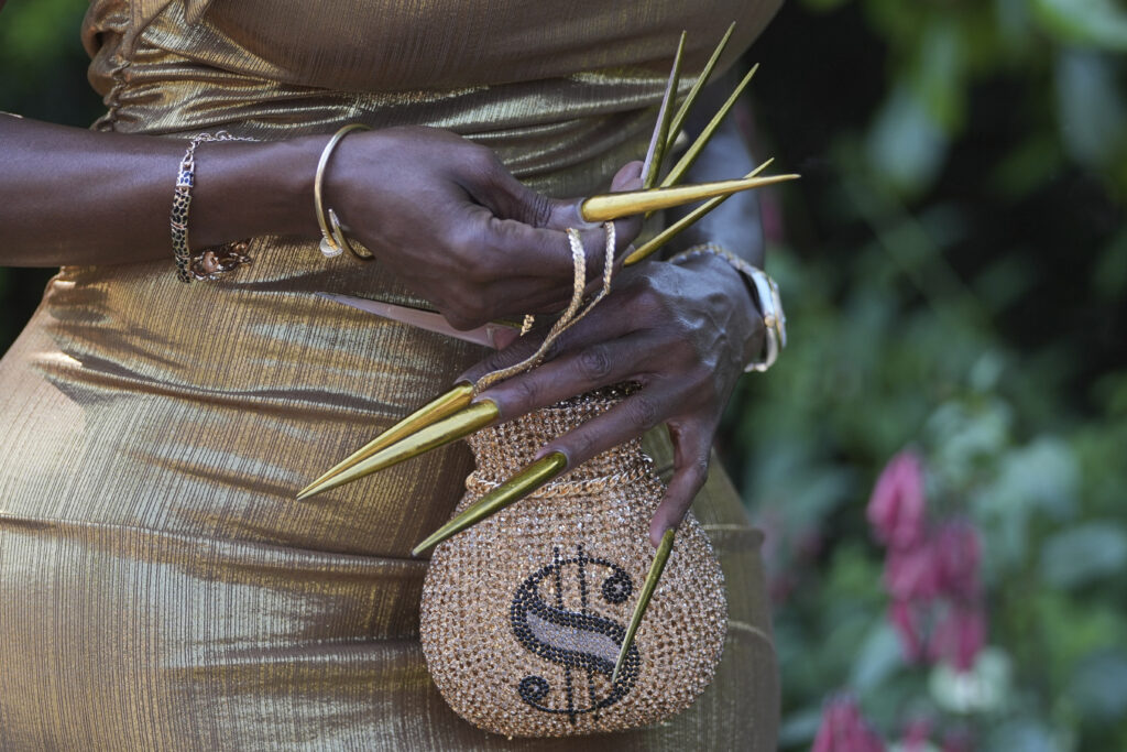 A racegoer in a gold dress with long gold nails and a dollar symbol handbag poses for a photograph on the third day of the Royal Ascot horse race meeting, traditionally called Ladies Day, at Ascot, England, Thursday, June 19, 2025.