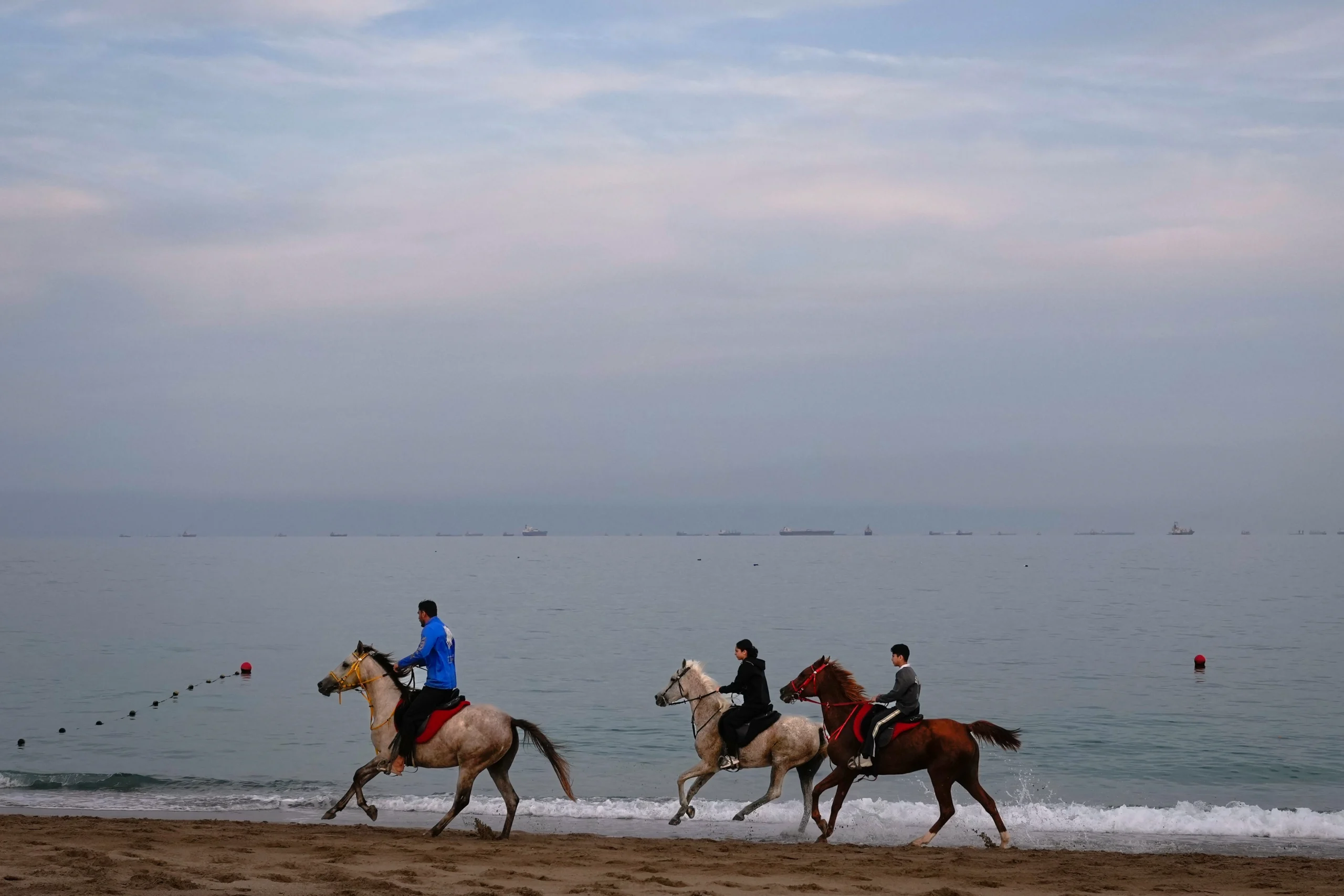 STRAIT OF HORMUZ — The eyes of the world are on this strip of water: Riders gallop along a beach as oil tankers and cargo ships line up in the Strait of Hormuz near Khor Fakkan, United Arab Emirates, Wednesday, March 11, 2026. Photo: Altaf Qadri/AP